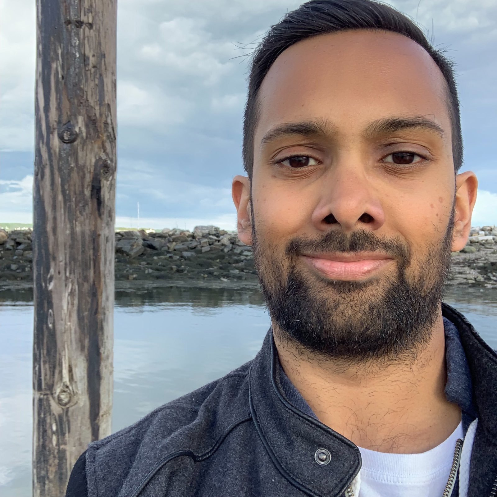 Ashik has light brown skin, short black hair, dark eyes and and a closely cropped black beard. he is smiling in front of a calm bay or pier. 
