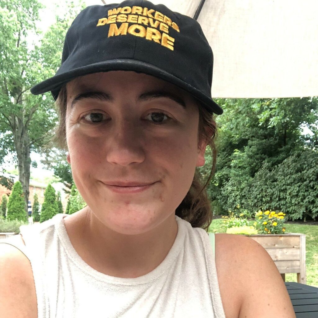 Cara has brown eyes and fair skin, her reddish brown hair is tied back and she is wearing a black cap that has "workers deserve more" embroidered in yellow