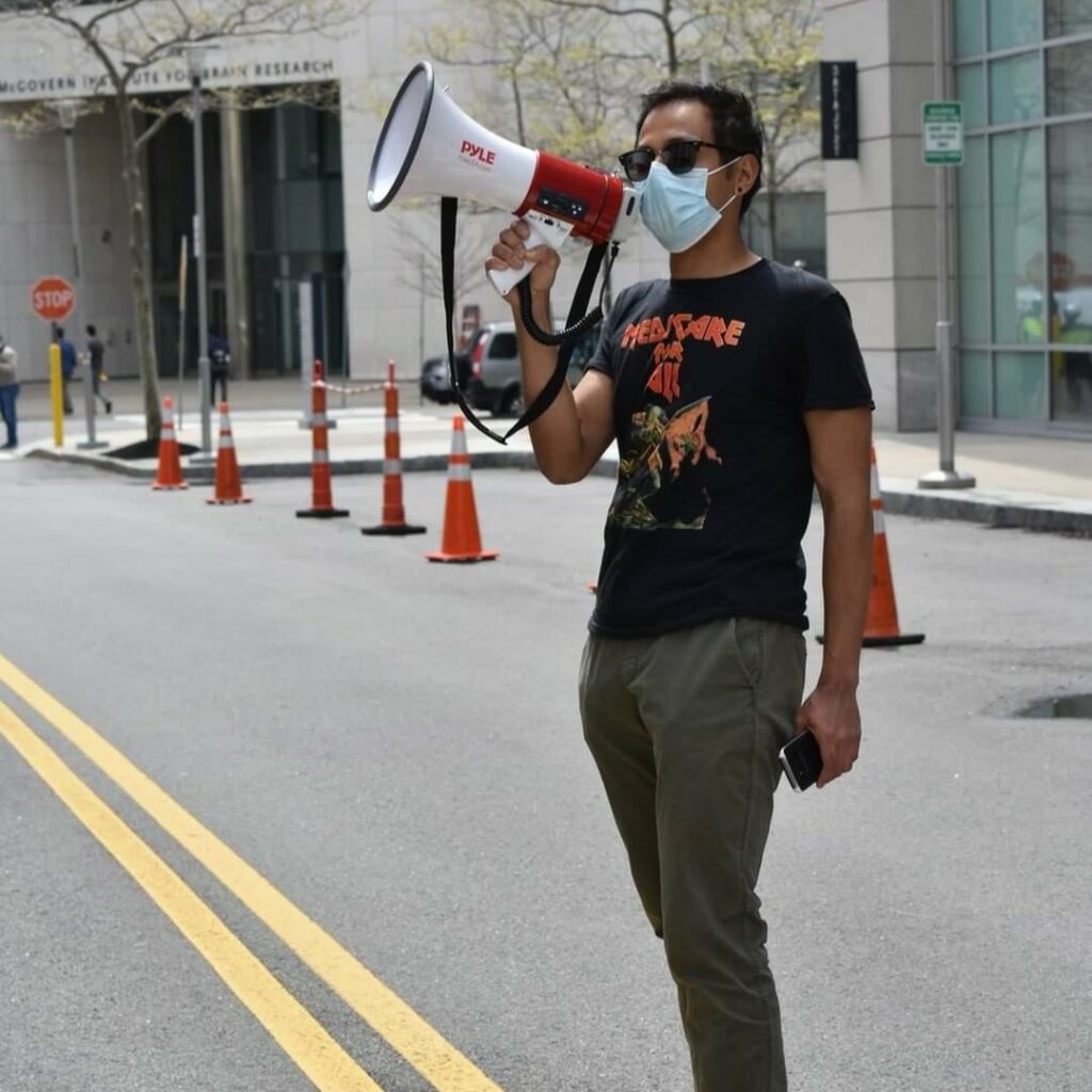 Nafis has brown skin and short, black hair. he is seen standing in a city street holding a bullhorn to his face while wearing sunglasses and a paper covid mask.