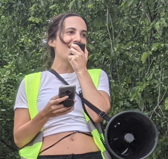 Nicole has fair skin and long dark hair. She is speaking into a bullhorn microphone and is wearing a yellow safety vest. There is foliage behind her.