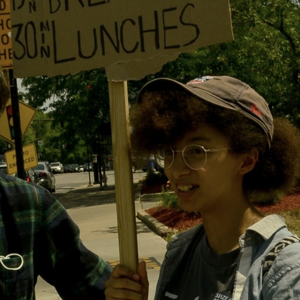 Soliel has dark olive skin and kinky curly brown hair, she sports thin silver glasses and a brown cap. she is outside holding a sign that says "honk for 15 min breaks + 30 min lunches"